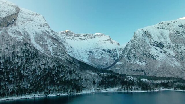 aerial footage showcasing the serene winter landscape of a Norwegian fjord, possibly Eresfjord or Eikesdalen. Snow covered mountains rise dramatically from the tranquil waters, surrounded by a dense, 