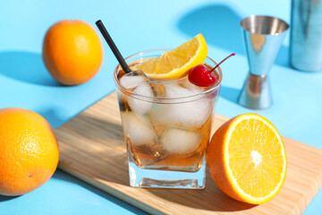 Glass of tasty Old Fashioned cocktail with cherry and fresh oranges on blue background