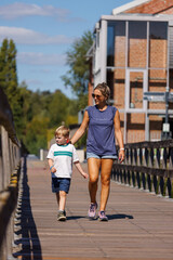 Obraz premium Mother and son walking hand in hand on wooden bridge in summer, casual family lifestyle outdoors near modern building