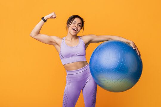 Workout for weight loss. Positive african american slim woman in sportswear holding fitness ball and showing biceps, isolated on yellow background, studio shot. Body care and sporty lifestyle concept - Powered by Adobe