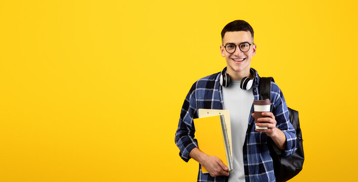 Happy male student with notebooks, backpack and takeaway coffee looking at camera and smiling on orange studio background. Cheerful young guy participating in educational program