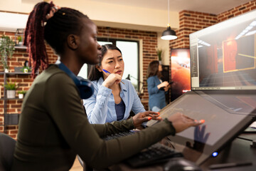 Focused woman manager closely reviewing the editing progress of black employee seated modern workstation. Asian team leader with a pen in hand, looking at computer screen while female designer works.