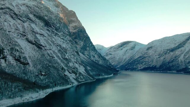Aerial footage capturing the serene beauty of a snow covered fjord in Norway during winter, possibly Eikesdalen or Eresfjord. Steep, majestic mountains blanketed in white snow rise dramatically from t