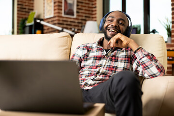 Black entrepreneur wearing headphones laughs at hilarious film on laptop, enjoying lighthearted...