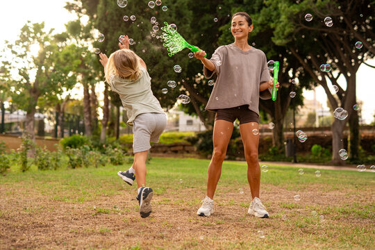 Smiling mother creating soap bubbles with a wand while her happy young son reaches to catch them, joyful family outdoor activity in the park on a summer day - Powered by Adobe