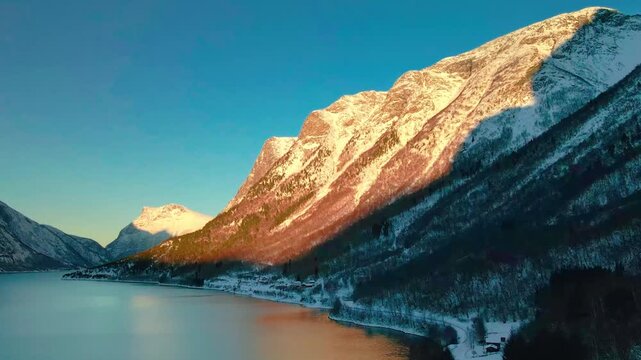  This drone footage shows the serene beauty of a tranquil Norwegian fjord at sunset, with the last rays of golden light illuminating the snow covered peaks and reflecting in the calm waters.