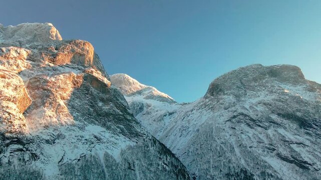 This drone footage captures the serene beauty of snow covered mountains in Norway, possibly within the stunning landscapes of Eresfjord or Eikesdalen, illuminated by the warm, golden light of dawn or 