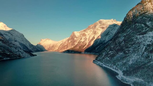 The drone shot of winter landscape footage capturing the serene beauty of a Norwegian fjord, potentially Eikesdalen or Eresfjord, with snow-capped mountains reflecting in the calm waters under a clear