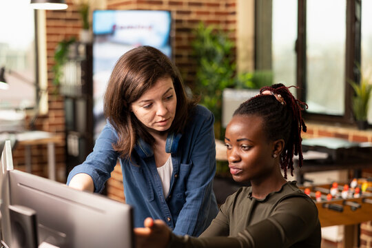Caucasian manager attentively listening as black woman shares progress updates, gesturing to computer screen. Closeup of female coworkers collaborating on a startup project, discussing creative ideas.