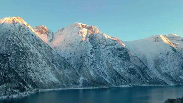 Aerial footage capturing the serene beauty of the snow capped alpine mountains surrounding a tranquil lake in Eresfjord, Norway, bathed in the warm glow of the golden hour.