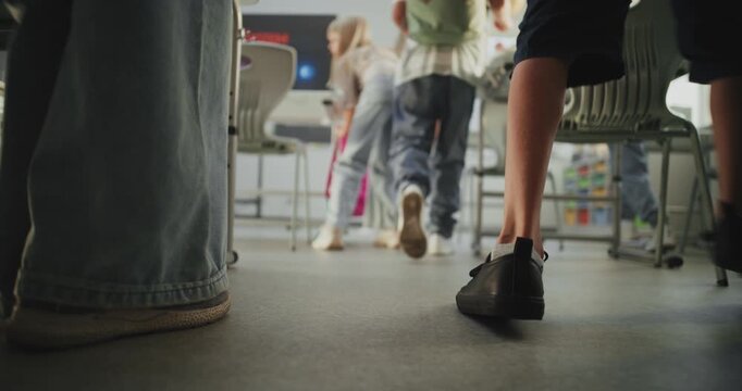 Knee Level Shot of Elementary School Children Entering Modern Classroom, Unpacking Their Backpacks and Laying Out School Supplies on Desks. Female Teacher Greeting Young Boys and Girls. Slow Motion.