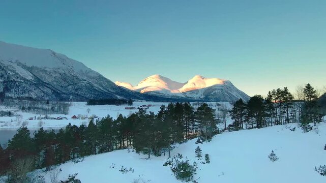 aerial footage captures the serene beauty of a winter sunrise illuminating snow covered mountains and evergreen forests in either Eresfjord or Eikesdalen, Norway.