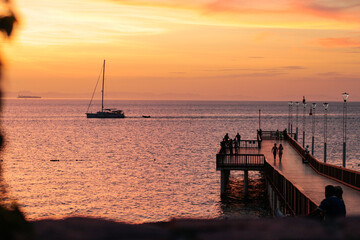 Fototapeta premium A serene golden sunset over the pier at Corumuel Beach, Baja California Sur, Mexico, with a sailboat on the horizon and calm ocean waters during golden Hour.