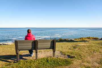 Fototapeta premium Woman enjoying a magnificent seascape while sitting on a bench on the top of a cliff on a clear autumn day. Pemaquid Point, ME, USA.