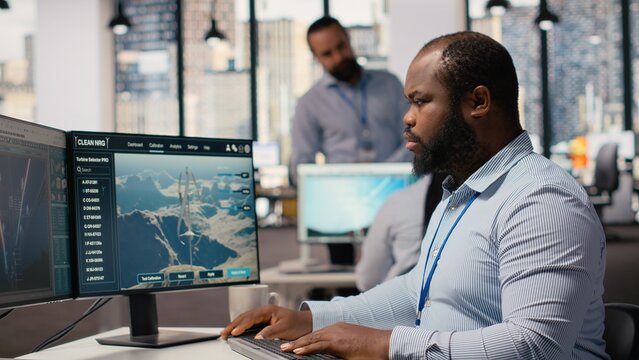 Employee using electric grid optimization tools with wind farm layout modeling software to enhance clean energy systems. Engineer doing power lines and futuristic wind turbine simulations, camera B