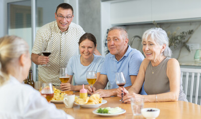 Happy senior couple, man and woman, with adult son and daughter gathering with family friends at cozy kitchen table, chatting cheerfully over drinks with snacks