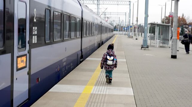Small child in winter jacket with penguin backpack walking alone on empty train station platform next to passenger train. Family travel, public transportation and childhood independence journey. 