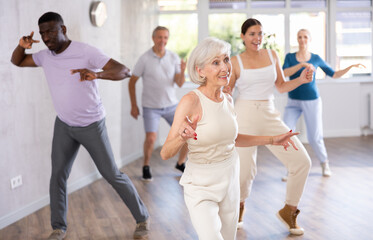 Cheerful sporty senior lady enjoying active dancing during group training in dance studio, practicing playful twist