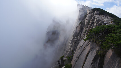 Misty clouds envelop a dramatic, rocky mountain peak, revealing rugged cliffs and sparse green vegetation
