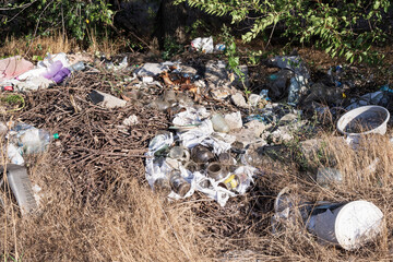 Pile of garbage in nature, including plastic bottles, bags, broken glass, and construction debris among dry grass and trees. Environmental pollution, waste management problem.