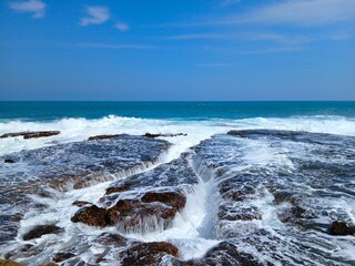 Waves Crashing into Rocky Holes on Coastal Shore with Blue Sky Horizon. Powerful ocean waves breaking into rocky gaps with foamy surf and scenic horizon view.