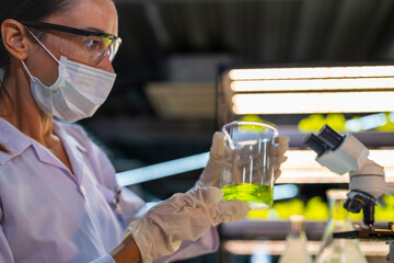 A scientist wearing a mask and gloves works in a lab, examining a green liquid in a beaker and using a microscope. The lab setup is well-lit, reflecting a focus on research, science, and experimentati