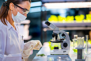 A scientist wearing a mask and gloves works in a lab, examining a green liquid in a beaker and...