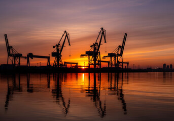 Obraz premium Silhouetted Industrial Cranes at Port During Vibrant Sunset Reflection
