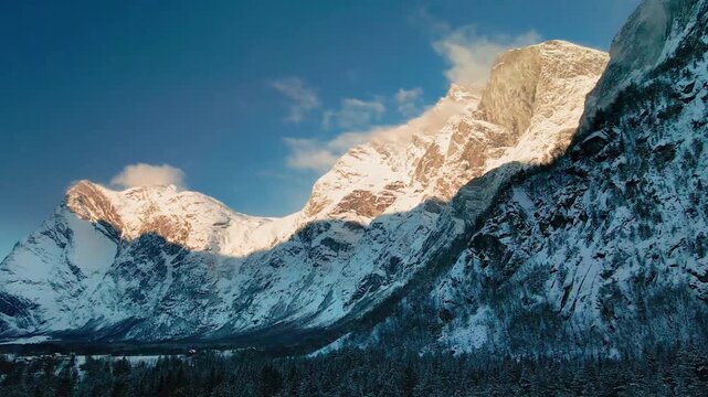 The drone footage capturing the iconic Trollveggen Troll Wall in Norway, bathed in the golden light of winter sunrise or sunset. The snow-covered peaks and dramatic rock faces stand tall against a cle