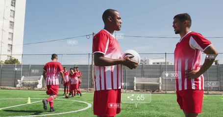 Holding soccer ball, mid adult male player facing teammate on artificial turf by fence, with cones