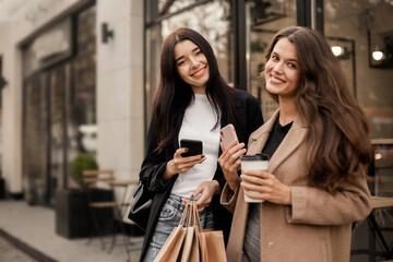 Two smiling women friends dressed in beige coat and jacket drinking coffee and using smartphone or phone, holding paper bags with purchases in hand after shopping walking along streets of autumn city.
