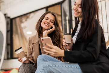 Two young women friends dressed in beige coat and jacket drinking coffee, having fun and chatting together and using smartphone sitting against the background of cafe in autumn seasons.