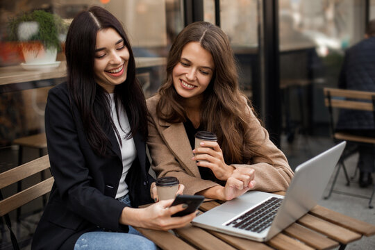 Two young cheerful women friends sitting in a street cafe, drinking coffee and using a smartphone and laptop for work or online shopping. Online purchases on Black Friday or Cyber ​​Monday.