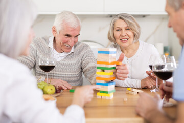 Two married couples are having fun in cozy home atmosphere, playing building tower with wooden blocks game. Senior participant is removing wooden block bricks from tower and others are watching
