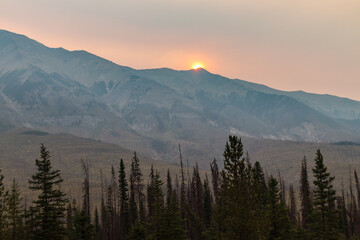 Obraz premium Golden sun sets behind rugged mountain peaks, casting a warm glow over the forested valley below