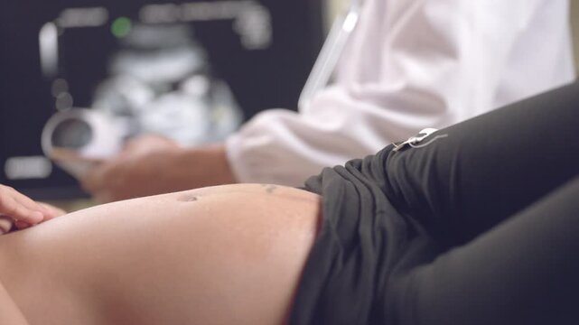 Medical professional using ultrasound device to examine pregnant belly while fetal image appears on screen.