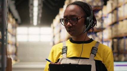 Call center female employee assisting clients in a storehouse depot, checking labels on packages and shelving units. Young adult resolving problems and delays related to delivery. Camera B.