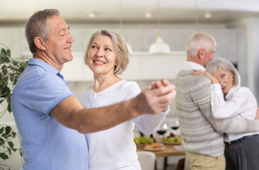 Obraz premium Senior men and women doing couple dancing during house party. Two married couples pensioners enjoy move to beat of lounge music in cozy homely atmosphere.