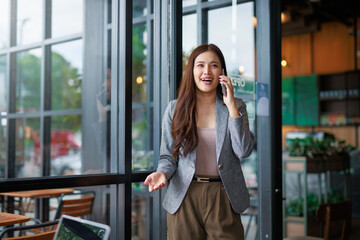 Asian businesswoman leaving a cafe, talking on her phone, smiling brightly and gesturing with her hand, exuding confidence and positivity