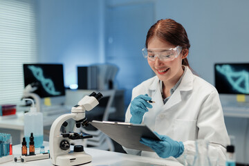 Asian female scientist smiling and taking notes on clipboard, working with microscope and dna structure on computer screen in modern laboratory