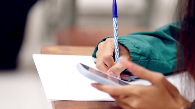 High school student multitasking with smartphone and schoolwork at the same time, writing on paper while using mobile device.