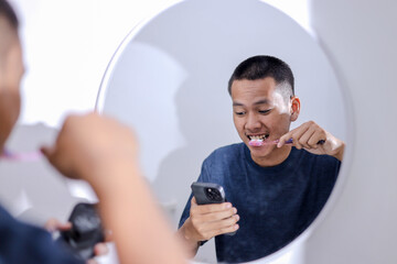 Happy Asian Man Using Smartphone While Brushing Teeth in Bathroom