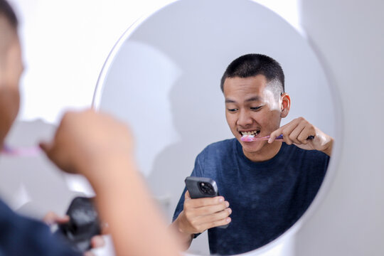 Happy Asian Man Using Smartphone While Brushing Teeth in Bathroom