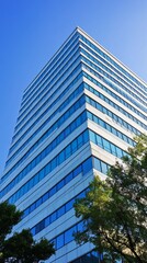 Obraz premium Low-angle view of a modern, tall building with blue windows and light gray panels, set against a clear blue sky, with some greenery at the base