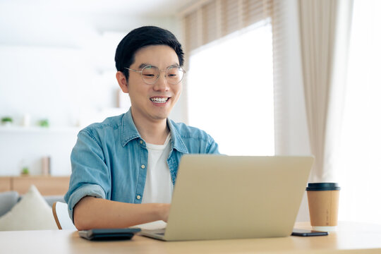 Happy young Asian man working on laptop in living room. Freelance worker male sitting at desk writing notes while watching webinar, studying online