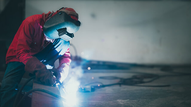 A welder in a red jacket and protective mask focuses intently on a metal piece in a workshop. Sparks fly as he works, illuminating the space with bright flashes during the evening