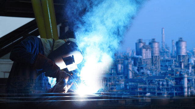A worker conducts welding at an industrial facility during twilight. Bright sparks fly as the welder focuses on his task, surrounded by machinery and smoke