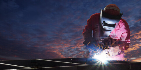 A welder in protective gear focuses on his work, creating sparks as he welds metal pieces together at sunset. The dramatic sky adds to the intensity of the moment