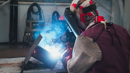 A welder works meticulously on a steel piece, sparks flying as the torch ignites. The dim workshop highlights the bright flame, showcasing craftsmanship and dedication