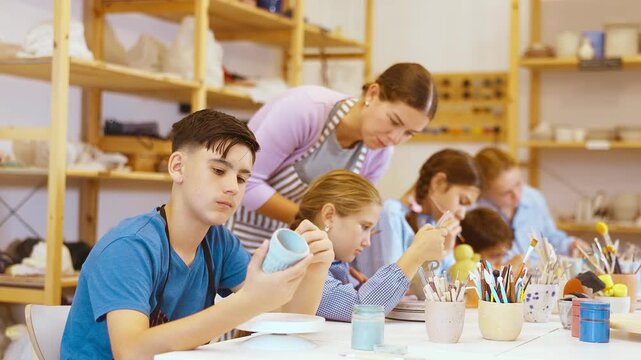 Children and teenagers sit at a table and paint ceramics at the workshop. Teacher shows how to paint a clay product after firing